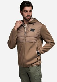 Ljusbrun zip-up jacka med en texturerad, ribbad kropp och slät, vadderad bröstparti. Har en huva och svart logotypaccent. Bärs över olivgröna byxor.