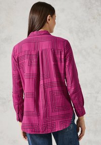 Camicia fucsia con bottoni, a righe geometriche nere, maniche lunghe con polsini a bottoni e una piega sul retro. Tessuto morbido e testurizzato.