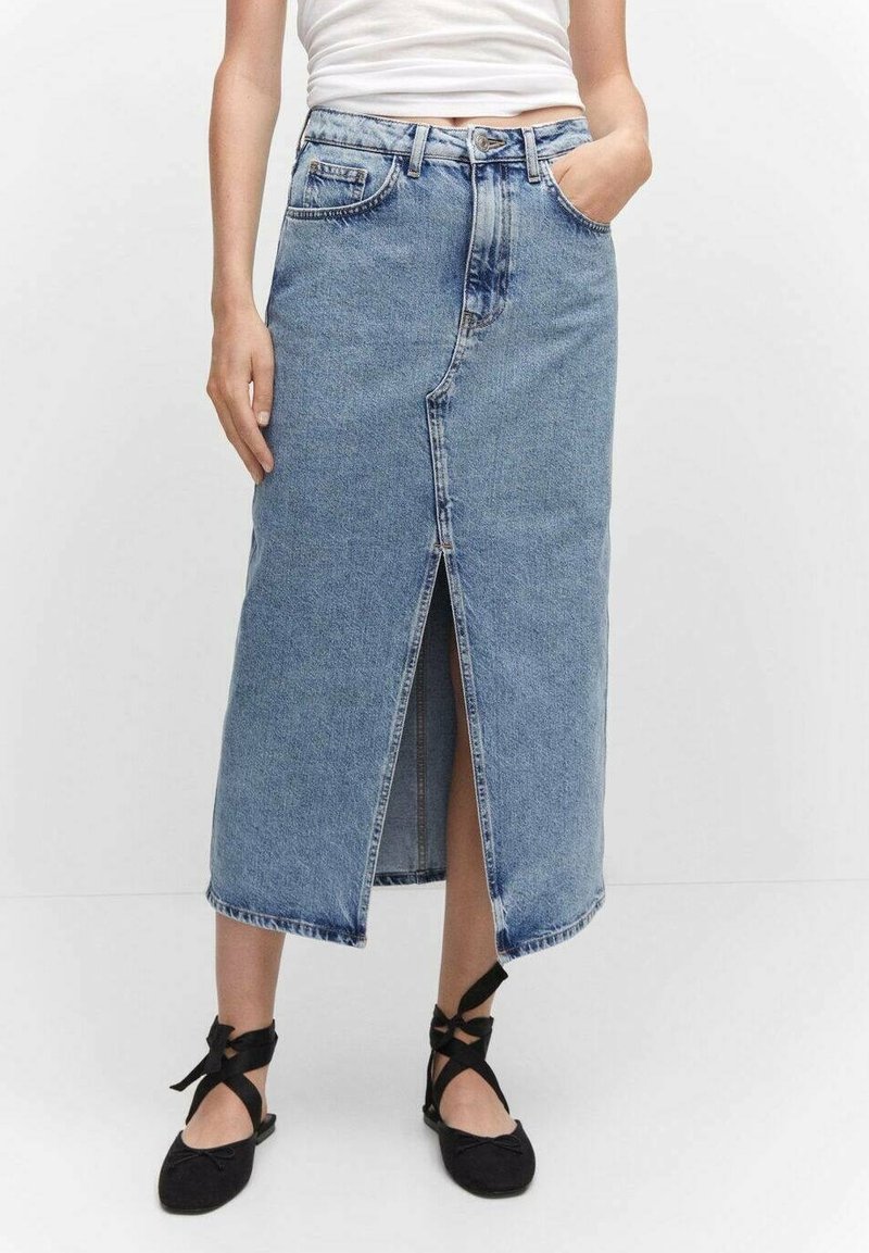 Personne portant une jupe midi en jean bleu avec une fente sur le devant, un haut blanc sans manches, des ballerines noires avec des rubans autour de la cheville, debout contre un mur blanc.