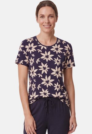 Smiling woman wearing a navy blue t-shirt with large beige flower patterns and dark drawstring pants against a white background.