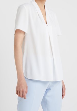 Vrouw draagt een losse witte korte mouwen blouse met V-hals, ingestopt in lichtblauwe broek tegen een effen achtergrond.