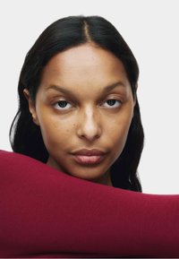 Zbliżenie portretowe na neutralnym tle, przedstawiające osobę z gładką skórą, ciemnymi włosami i bordowym materiałem zarzuconym na ramię.