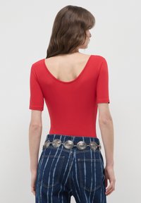 Donna con capelli castani ondulati indossa un top rosso con scollatura a barchetta, jeans a vita alta blu a righe e una cintura metallica argentata con design circolari.