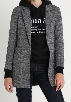 Personne portant un blazer gris texturé sur un sweat à capuche noir avec texte blanc et un jean bleu foncé, debout devant un fond uni.