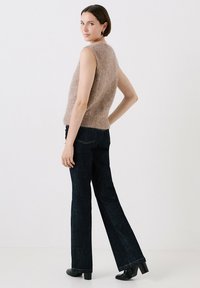 Mouwloze, lichtbruine gebreide trui met een zachte textuur, gecombineerd met donkere uitlopende jeans. Zwarte hakken, staande tegen een neutrale achtergrond.
