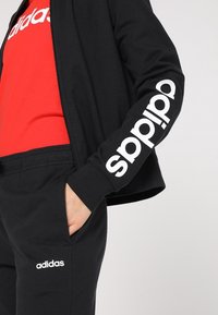 Personne portant une veste Adidas noire avec logo sur la manche, un t-shirt Adidas rouge et un pantalon Adidas noir avec logo près de la poche, main dans la poche.
