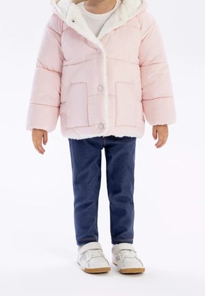 Bambino che indossa una giacca imbottita rosa chiaro con fodera in pile bianco, jeans blu e sneakers bianche, in piedi su uno sfondo bianco.