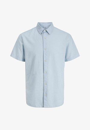 Jongen met een rode pet, lichtblauw shirt met korte mouwen over een marineblauw T-shirt met print, knielange spijkerbroek, witte sokken en bruine schoenen, lachend met een zijwaartse blik.