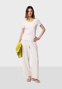Witte t-shirt met korte mouwen en een heldere gele halslijn, gecombineerd met crèmekleurige wijdvallende broek. Het model houdt een groene laag erop en draagt slippers.