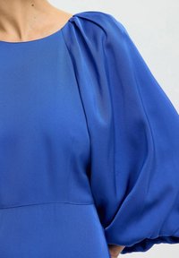 Blusa azul con mangas abullonadas y una textura suave y brillante. La tela cae elegantemente, destacando un escote redondeado y simple.