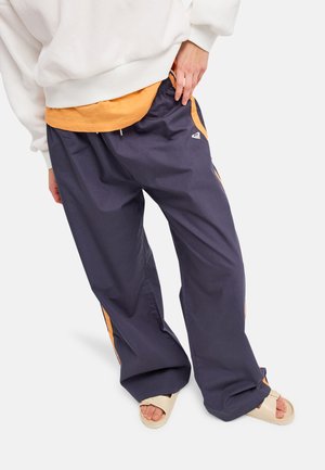 Persoon draagt losse marineblauwe trainingsbroek met oranje zijstrepen, beige slides, oranje shirt en witte trui, hand in de zak.