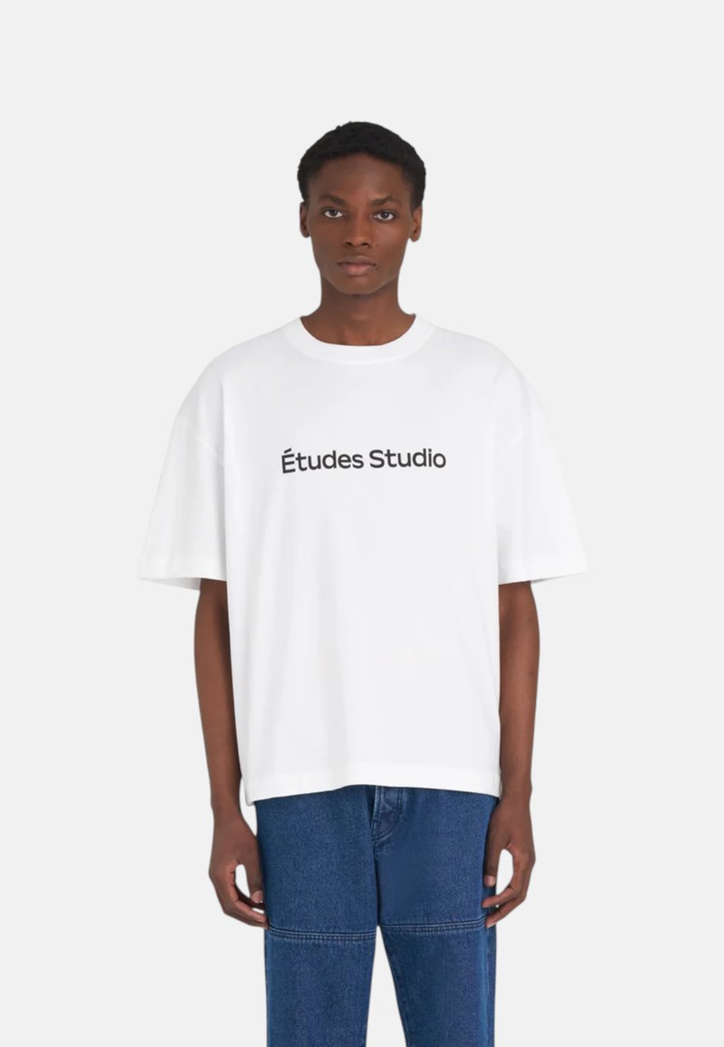 Personne portant un T-shirt blanc oversize avec le texte « Études Studio » et un jean bleu, debout devant un fond uni clair.