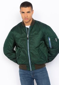 Giacca bomber verde con finitura satinata, zip frontale, polsini e orlo a costine, e tasca con patta sul braccio sinistro.