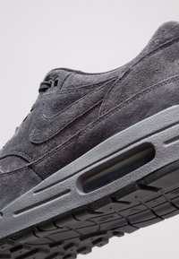 Šedá semišová teniska s texturovaným povrchem, obsahující viditelnou vzduchovou vrstvenou jednotku a odolným vzorem gumové podešve. Logo na boku.