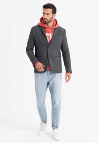 Grau blazer über einem korallenfarbenen Hoodie mit weißem Druck, kombiniert mit hellblauen Jeans und weißen Sneakers. Strukturiertes Design, lässige Mischung.