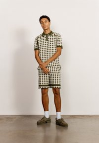 Grön och vit rutmönstrad pikétröja och matchande shorts med ribbade kragar. Modellen har på sig olivfärgade sneakers och vita strumpor, stående på en betongyta.