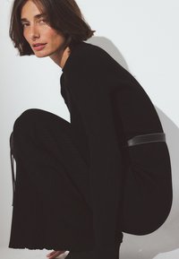 Črna rebrasta obleka s dolgimi rokavi in prilegajočo se silhueto, poudarjena s črnim usnjenim pasom. Tekstura je mehka in strukturirana.