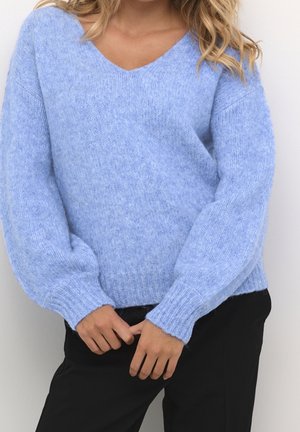 Femme portant un pull en tricot doux bleu clair avec de longues manches bouffantes et un pantalon noir, debout, les mains jointes devant.