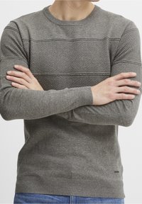 Uomo che indossa un maglione grigio a maglia strutturata con le braccia incrociate, jeans blu visibili in vita.