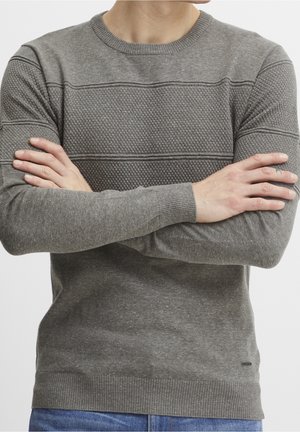 Mężczyzna w szarym teksturowanym swetrze z dzianiny, z założonymi na siebie ramionami, z niebieskimi dżinsami widocznymi w pasie.