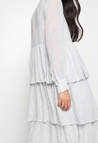 Vestido de capa azul claro en tejido transparente con un patrón sutil, mangas largas, puños abotonados y una silueta fluida.