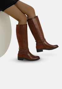 Botas de imitación de cuero marrón hasta la rodilla con cremallera lateral, correa decorativa y un tacón bajo cuadrado, presentando una combinación de texturas mate y brillantes.