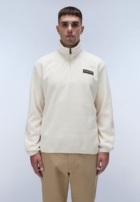 Cremefarvet fleece-pullover med høj hals, kvart-lynlås og et lille sort logo-patch på brystet. Løs pasform og blød tekstur.