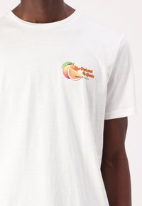 Λευκό βαμβακερό μπλουζάκι με γραφικό ροδάκινου που περιέχει κείμενο "Ο Μπαξέας της Ισπανίας" και "Ποιοτική Προϊόν" σε πορτοκαλί και πράσινες αποχρώσεις.