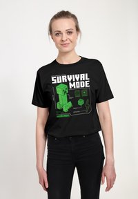 Czarny t-shirt z zielonym pikselowym wzorem postaci oraz napisem „SURVIVAL MODE” w pogrubionej białej czcionce, z elementami graficznymi związanymi z grą.