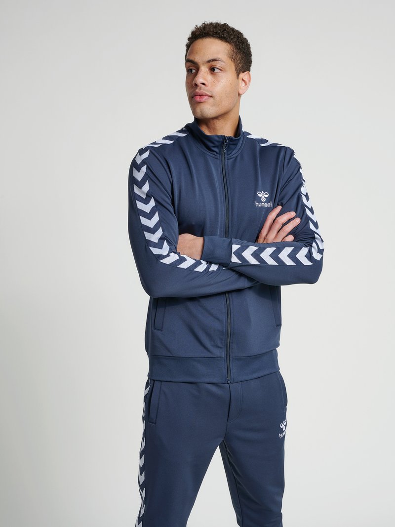 Junger Mann im dunkelblauen Hummel-Trainingsanzug mit weißen Chevron-Streifen, der mit verschränkten Armen dasteht und zur Seite schaut vor schlichtem Hintergrund.