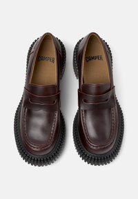 Brązowe skórzane mokasyny z zaokrąglonym noskiem, przeszyciami i kontrastującą czarną gumową podeszwą. Posiadają pasek na wierzchu.