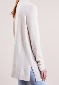 Pull en maille beige clair avec un col montant, un ourlet côtelé et des fentes sur les côtés. Texture douce, manches longues, conçu pour une coupe décontractée.