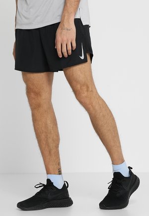 Uomo che indossa pantaloncini da corsa neri Nike, scarpe da ginnastica nere e calzini azzurri, in piedi con un piede leggermente avanti su sfondo bianco.