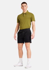 Polo olive avec deux boutons, associé à un short à cordon noir. Baskets montantes blanches en toile texturée.