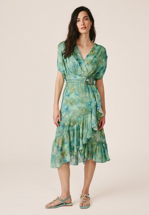 Kobieta ubrana w zielono-niebieską kwiatową sukienkę kopertową z falbankami i paskiem, w połączeniu z pasującymi płaskimi sandałami zdobionymi klejnotami, stojąca na jednolitym tle.