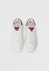 White sneakers with a smooth leather upper, maroon suede accents on the heel, and white laces. Features a rubber sole and logo on the tongue.