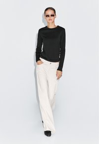 Sort langærmet top parret med løse hvide bukser med lommer. Accessories inkluderer smalle solbriller og ringe; baggrunden er neutral.