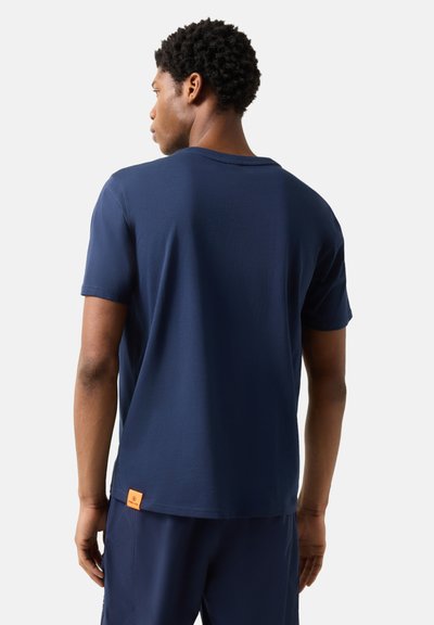 Homme de dos portant un t-shirt à manches courtes bleu marine et un short assorti bleu marine, sur un fond blanc uni.