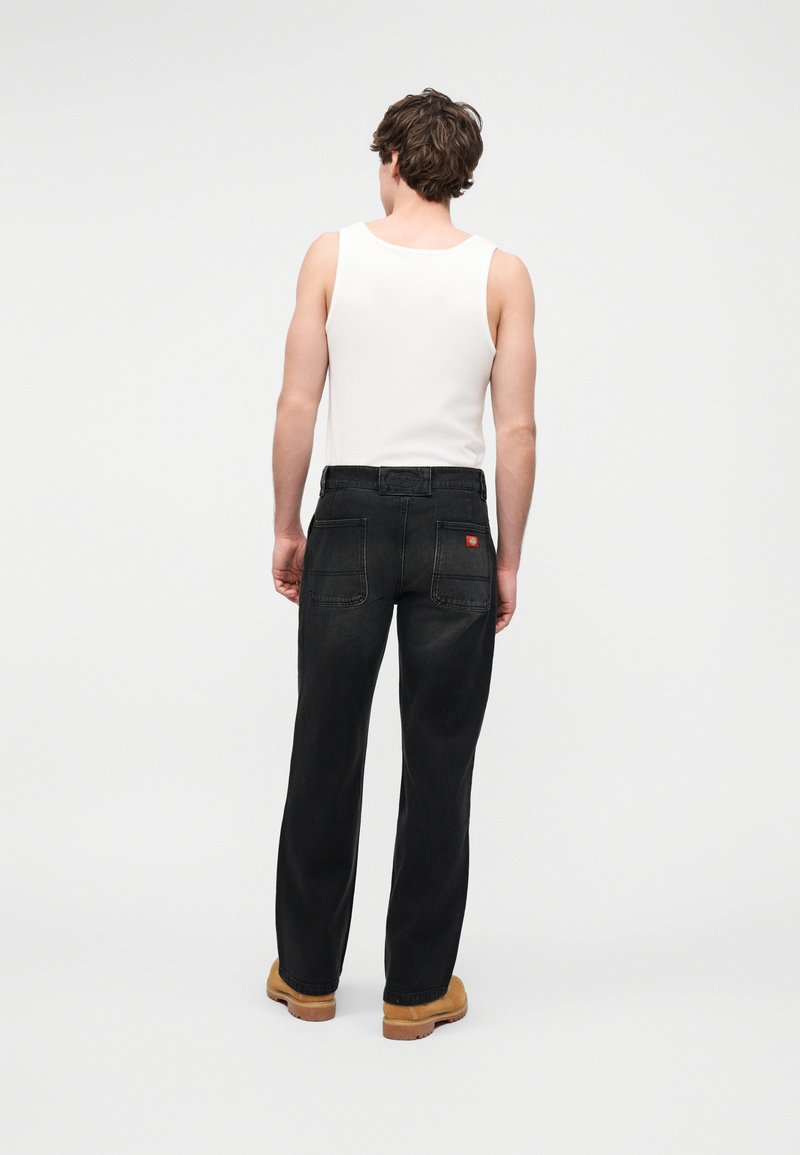 Man draagt een wit mouwloos hemd, zwarte spijkerbroek met achterzakken en beige werkschoenen, staat met de rug naar de camera op een effen achtergrond.