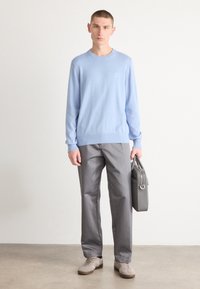 Hellblauer Strickpullover mit Rundhalsausschnitt, graue Hose und graue Tasche. Das Modell steht auf einer flachen Oberfläche; weiche Textur und moderne Designelemente.