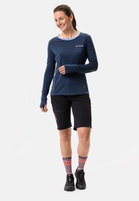 Camicia a maniche lunghe blu navy con scollatura azzurra, abbinata a pantaloncini neri. Calze a fantasia vivace e calzature nere completano l'outfit.