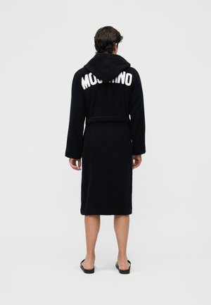 Άνδρας φοράει μαύρο μπουρνούζι με κουκούλα και το κείμενο "MOSCHINO" στην πλάτη, στέκεται ξυπόλυτος μπροστά σε μια απλή ανοιχτόχρωμη φόντο.