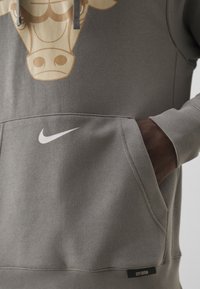 Main dans la poche d'un sweat à capuche gris Nike doté d'une poche kangourou, d'un logo swoosh blanc et d'un graphisme représentant une tête de taureau beige.