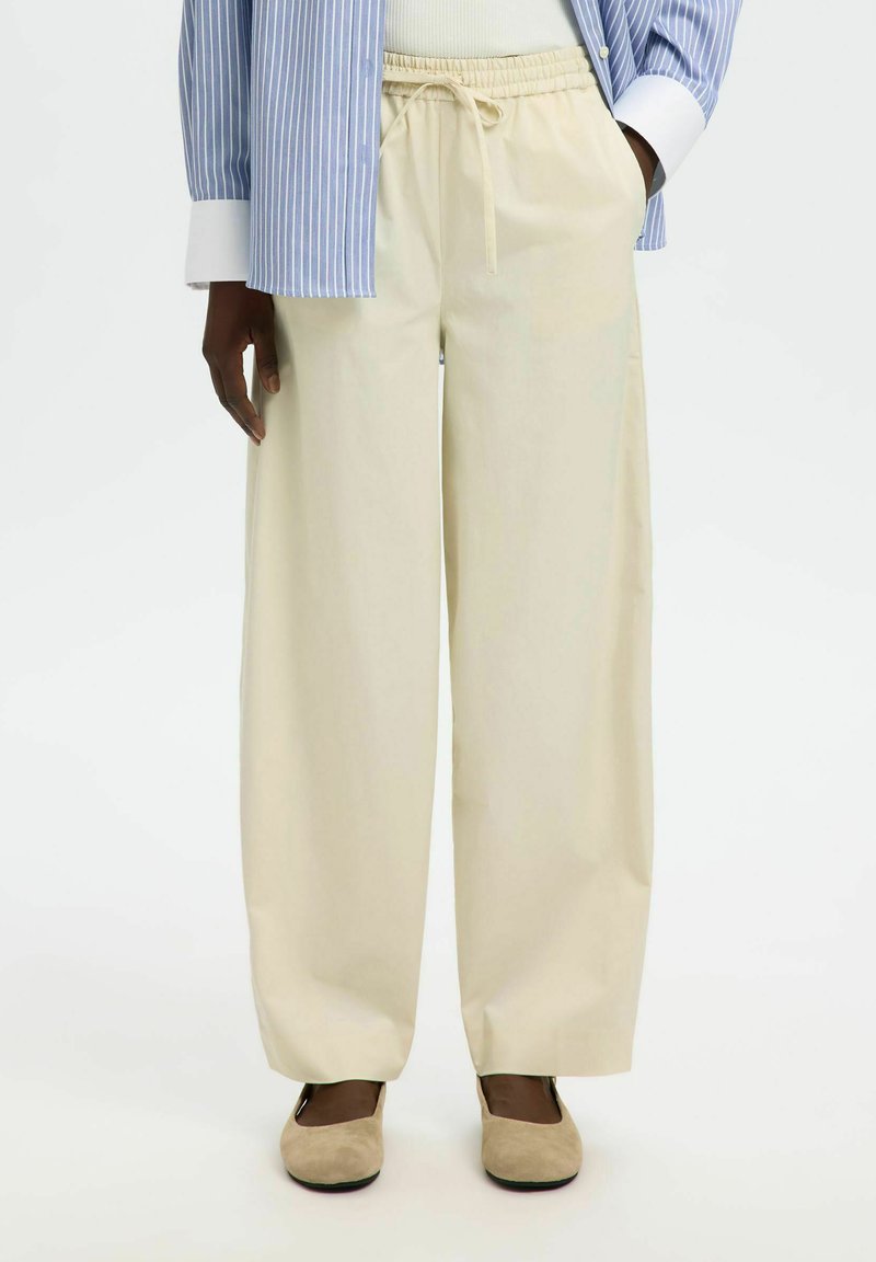 Personne portant un pantalon beige à jambes larges avec taille à cordon, des ballerines beiges et une chemise rayée bleu et blanc avec des poignets blancs.