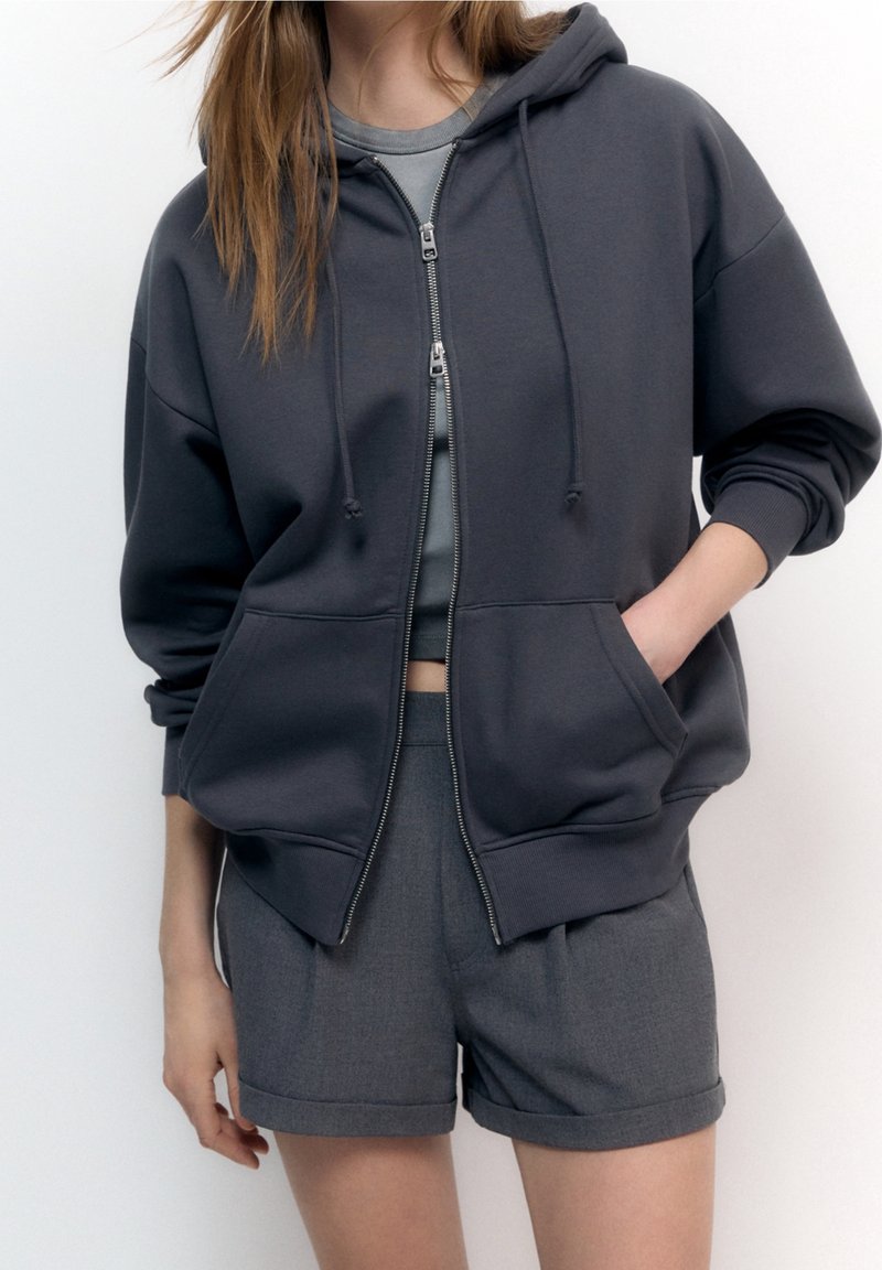 Jeune personne portant un sweat à capuche zippé gris foncé par-dessus un crop top gris et un short gris assorti avec ourlet roulé, debout devant un fond uni.
