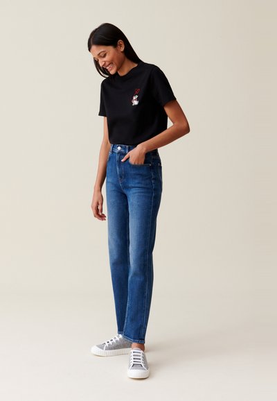 Femme souriante avec une main dans la poche, portant un t-shirt noir avec un design de chien et de cœur, un jean bleu et des baskets grises, sur un fond neutre.