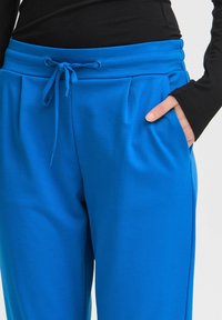 Modré běžecké kalhoty vyrobené z hladkého materiálu, s elastickým pasem na šnůrku, postranními kapsami a jemnými sklady na přední straně.