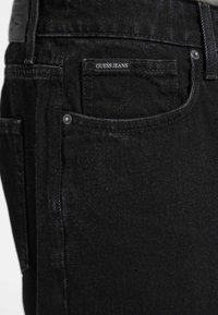 Jeans negri din denim cu o textură netedă, având un buzunar posterior cu un etichetă albă "GUESS JEANS" și accente din rivete metalice.