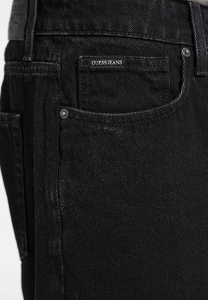 Jeans de mezclilla negra con una textura suave, que presentan un bolsillo trasero con una etiqueta blanca de "GUESS JEANS" y acentos de remaches metálicos.