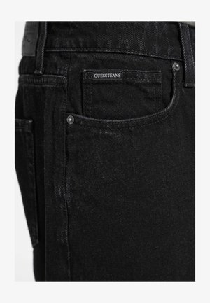 Fekete farmernadrág sima textúrával, hátul egy fehér "GUESS JEANS" címkével ellátott zsebből és fém szegecsekből álló részletekkel.